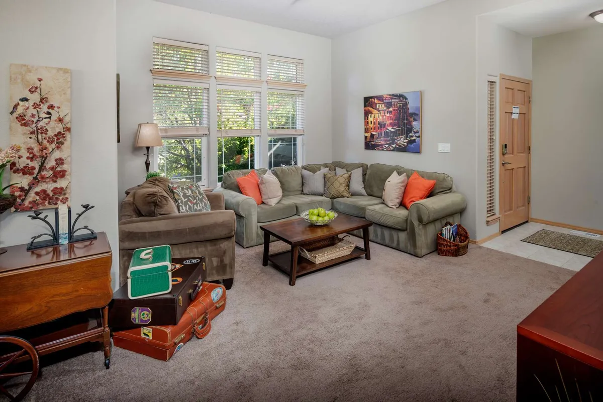 A similarly inviting living room with a plush sofa, coffee table, eye-catching artwork, and windows letting in natural light. A piece of luggage subtly signals the space's readiness for travelers and remote workers, while the arrangement supports both solo work and casual gatherings.