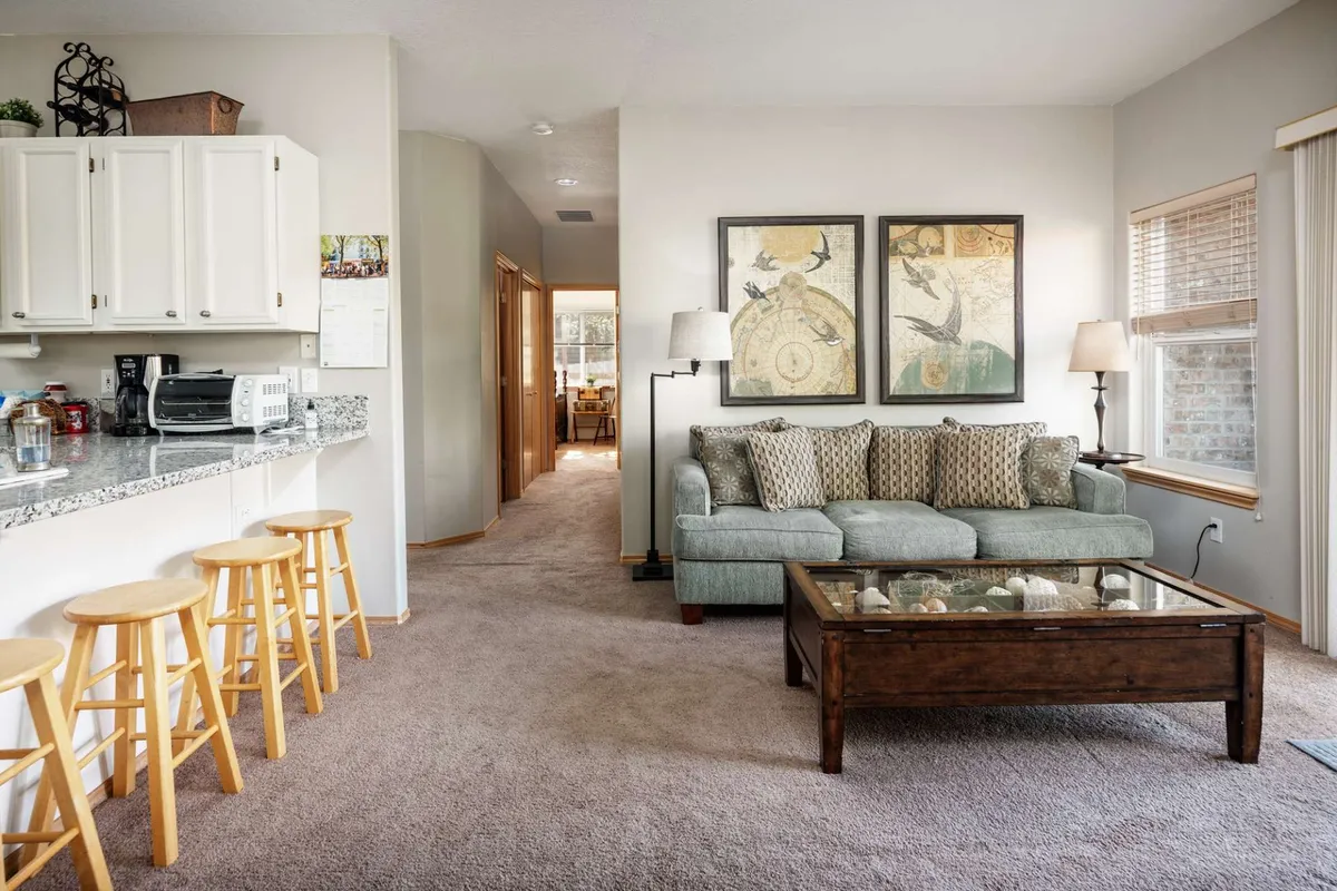 A welcoming living room featuring a sofa with patterned cushions, a glass coffee table, and a kitchen bar area with stools in the background. The layout encourages socializing, relaxing, or enjoying family activities in a single-level accessible setting. Framed wall art and warm lighting contribute to the inviting mood.