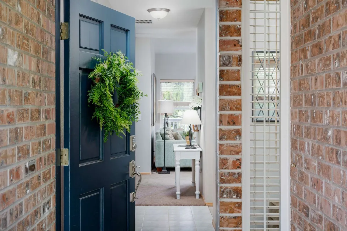 A welcoming entryway featuring a navy blue front door adorned with a green wreath, a table with a lamp, and a window letting in natural light. The scene has a bright, friendly atmosphere that hints at the transition between the indoors and the neighborhood just beyond.