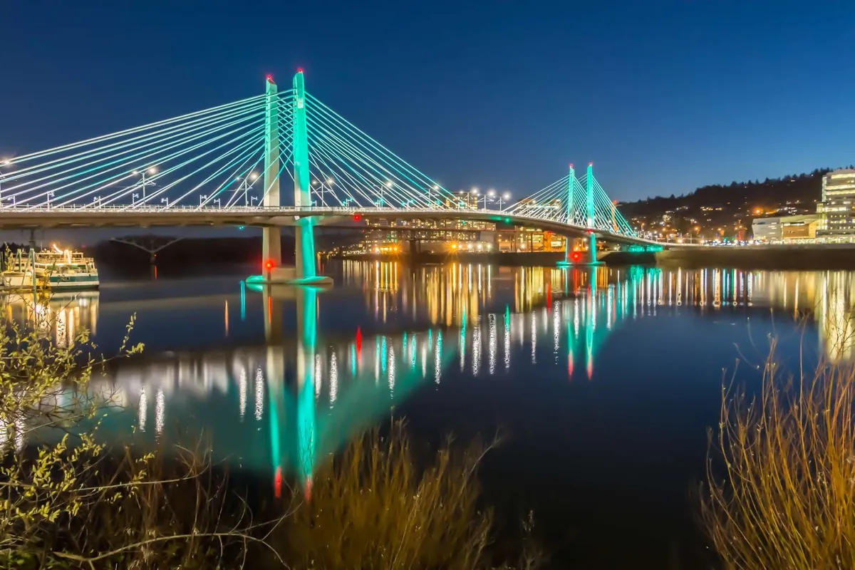 This image captures an illuminated bridge reflecting over tranquil waters, with a lively cityscape glowing in the background. The scene evokes a sense of exploration and the enjoyment of scenic surroundings, suggesting adventures beyond the retreat.