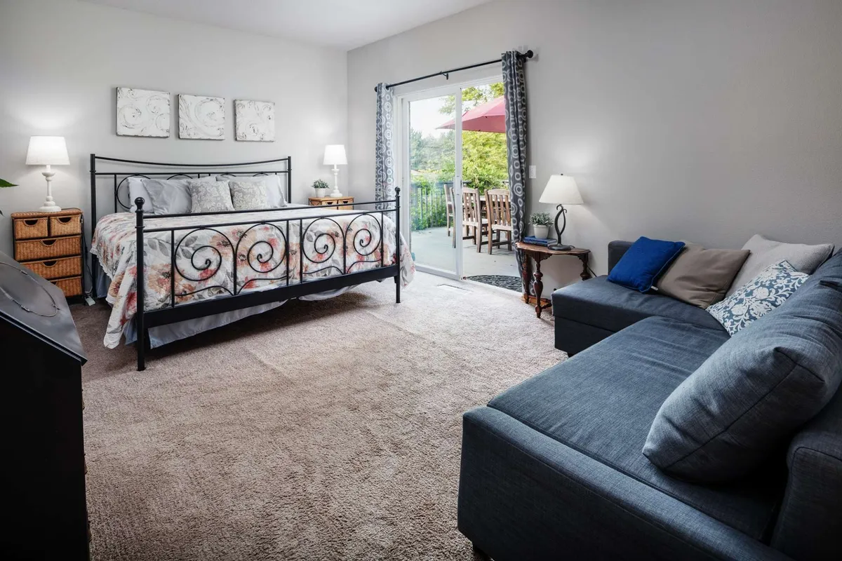 A serene bedroom featuring a bed with an ornate headboard, a sliding glass door opening to outdoor seating, and a cozy couch by the window. The room is filled with natural light and offers direct views of the surrounding greenery, creating a harmonious blend of indoor comfort and outdoor tranquility.