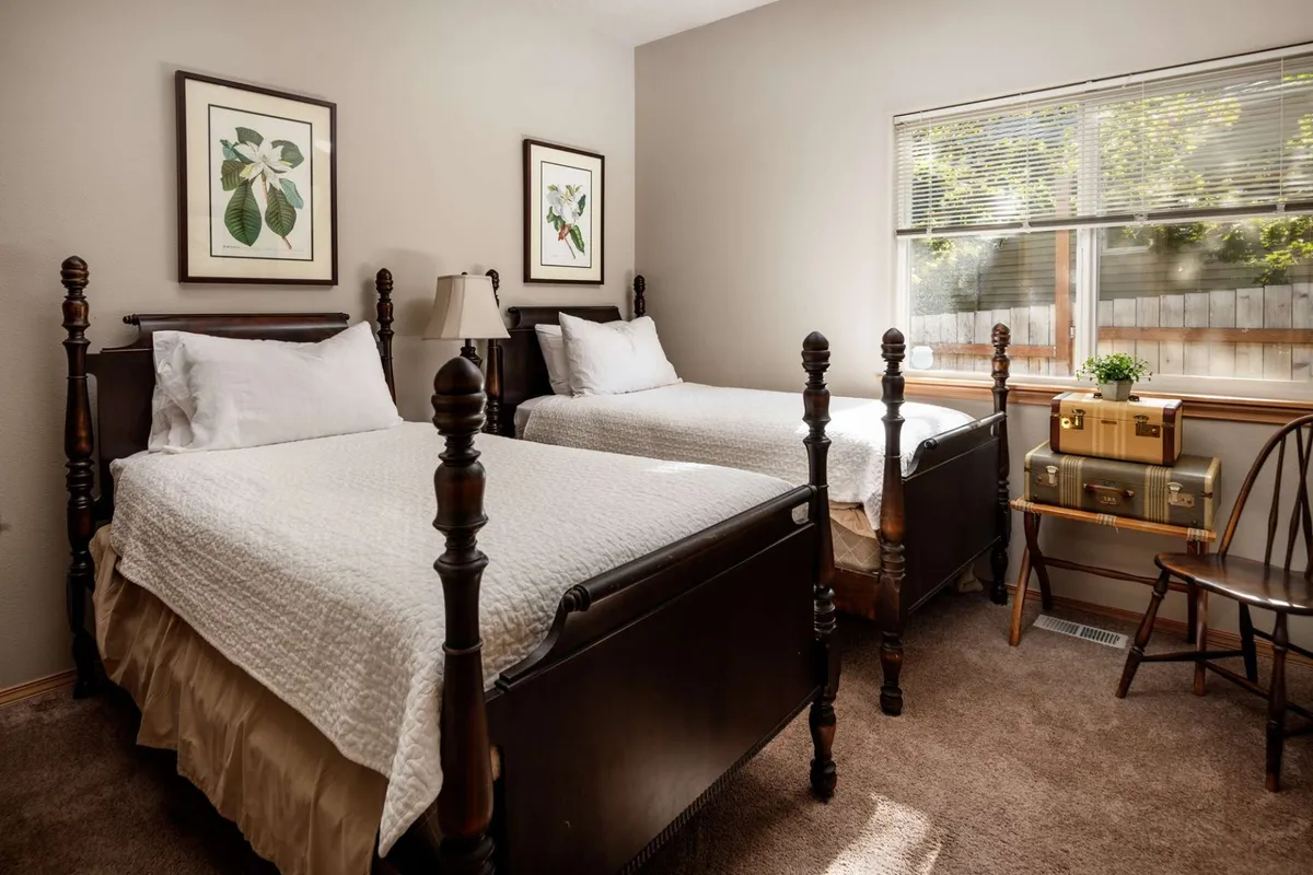 A cozy bedroom with two wooden beds featuring ornate headboards, framed botanical prints on the walls, and a small vintage desk. Morning light streams through the window, illuminating the inviting space ideal for children or multiple guests.