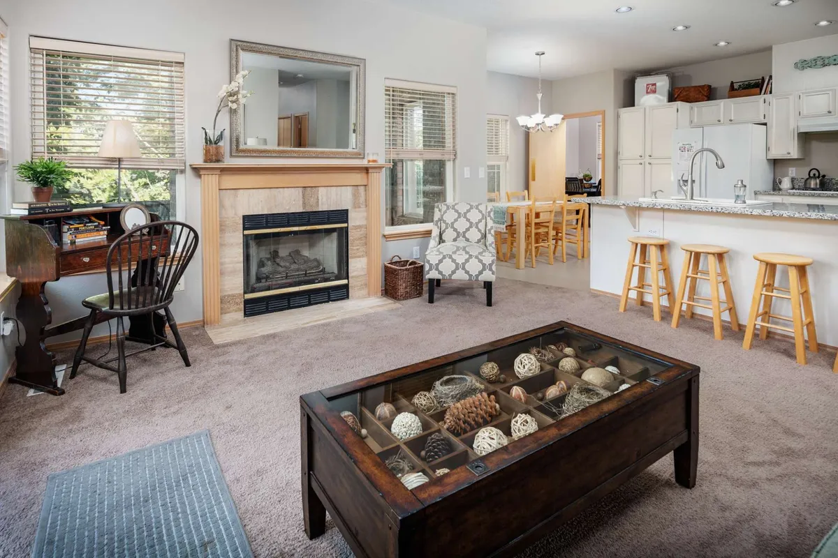 This image showcases a warm, welcoming living area anchored by a fireplace, with an inviting coffee table and comfortable armchair. The windows subtly hint at the greenery outside, suggesting proximity to nature even from within the home.