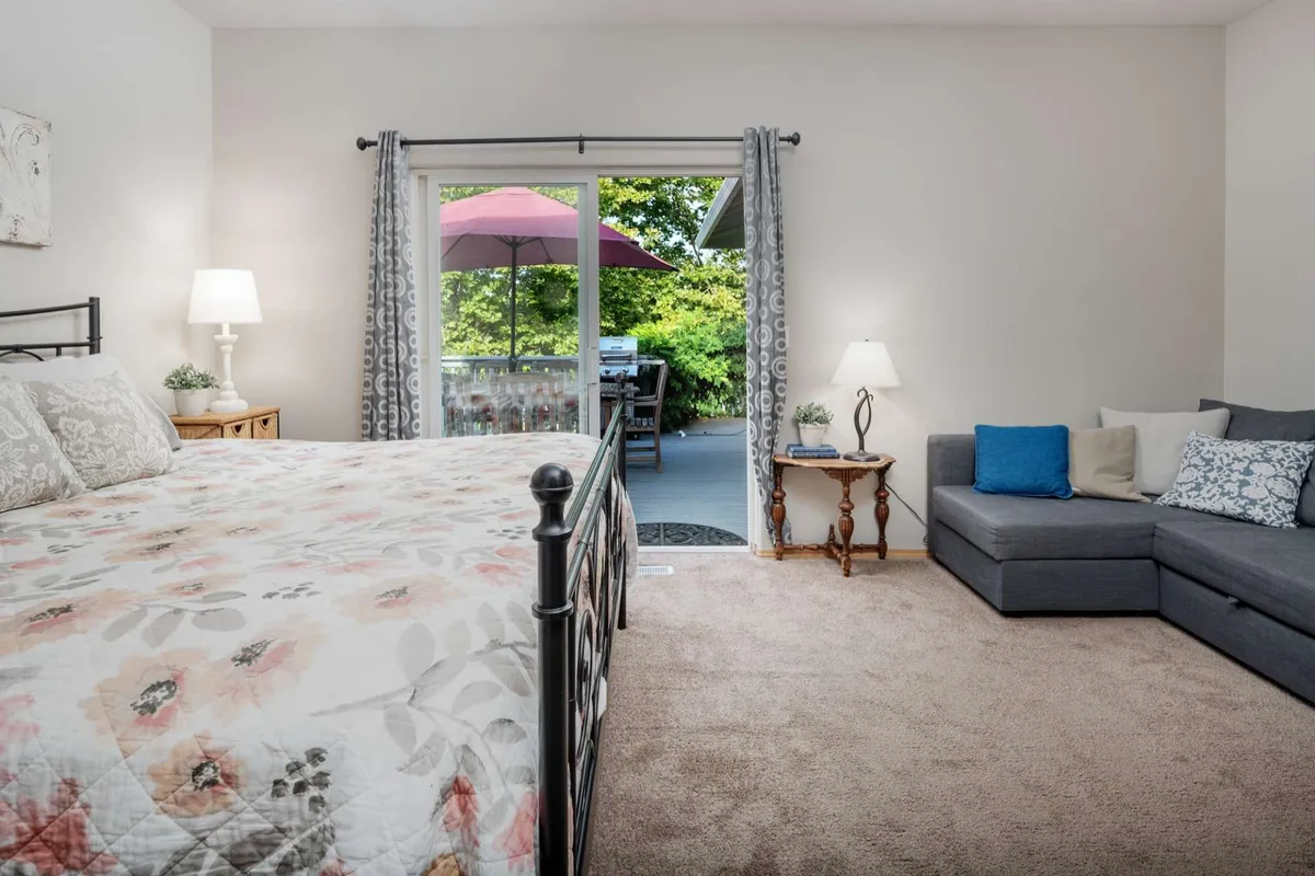 A serene living area that seamlessly blends a cozy sofa with colorful cushions and a bed with floral bedding, all bathed in natural light from a sliding glass door leading outdoors. This space offers versatile sleeping options and a comfortable setting for both socializing and rest, with easy access to the outdoors.