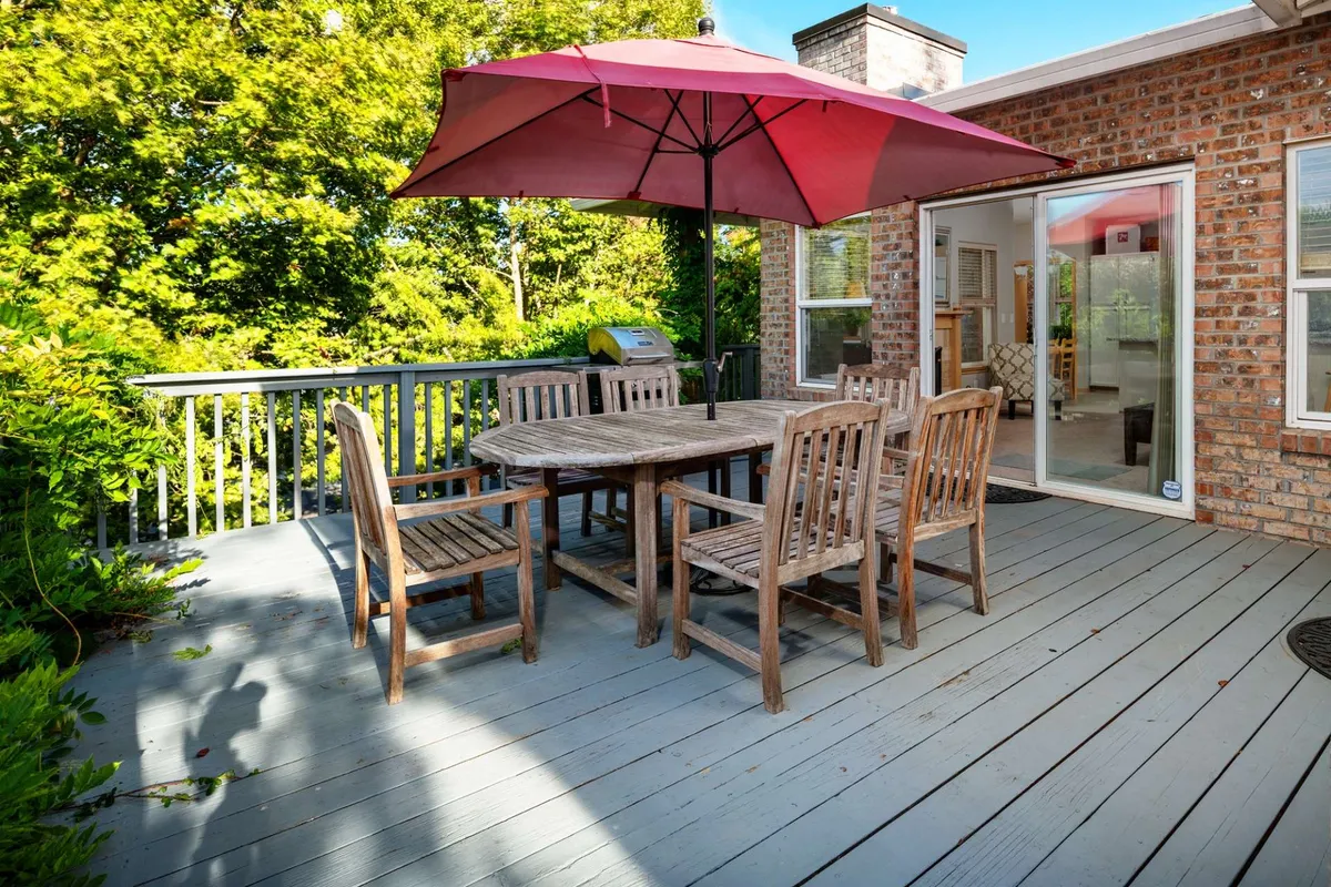 The image showcases the home's inviting backyard patio, featuring a sturdy wooden dining table shaded by a vibrant red umbrella, surrounded by lush green foliage. A sliding glass door leads directly from the house to the outdoor space, suggesting seamless indoor-outdoor living. The scene is bathed in cheerful daylight, hinting at opportunities for gatherings, barbecues, and relaxation in a tranquil, wooded setting.