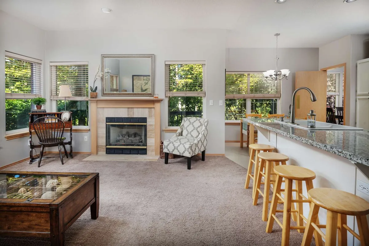 A spacious, light-filled living area with a granite countertop, stools, and a fireplace. The layout encourages gatherings and reflection, with windows that allow glimpses of the natural setting beyond.