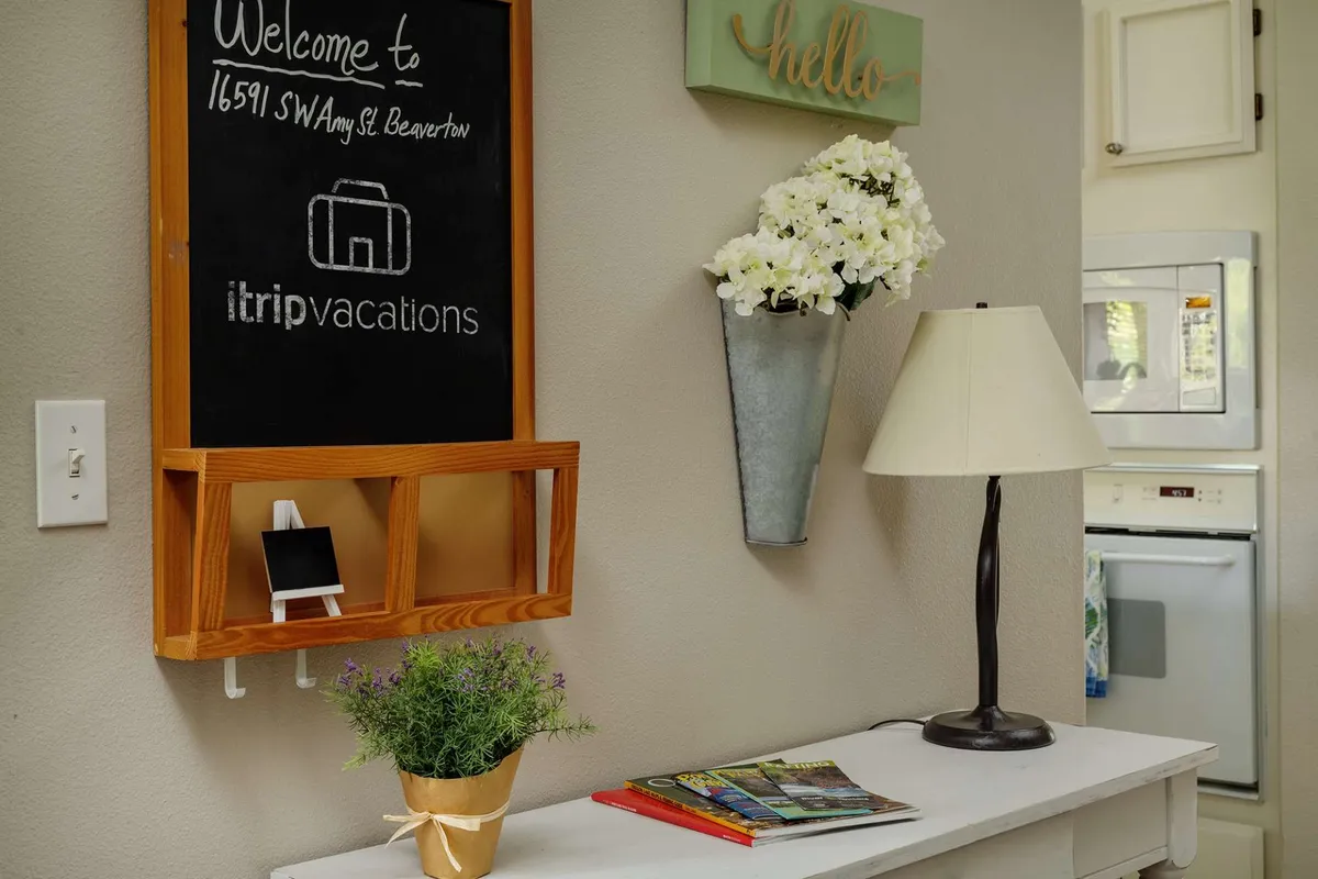 An inviting entry table with a welcome chalkboard, a fresh flower arrangement, a table lamp, and a stack of magazines. The scene is cheerful and hints at guests planning outings or discussing local adventures in a space designed for gathering.