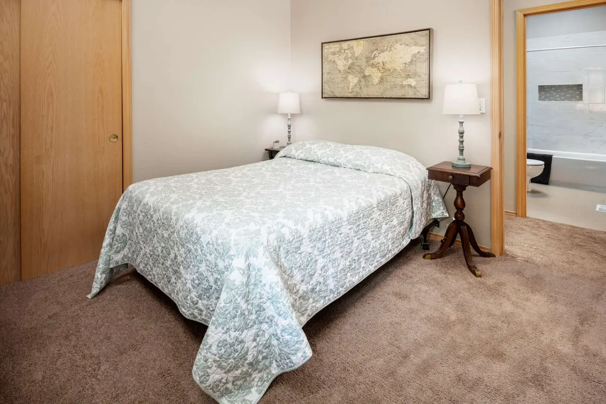 A tranquil bedroom with a bed dressed in a floral coverlet, nightstands with lamps on either side, and world map artwork above the headboard. Soft sunlight filters through the room, enhancing the restful and inviting mood.